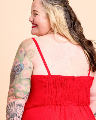 A woman modeling a bright red midi dress from the back, standing against a solid light beige studio background. This is the Wayland Dress sewing pattern from Cashmerette.