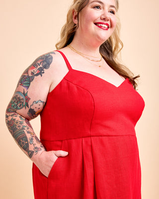 A woman with a tattooed arm wearing a bright red sleeveless jumpsuit with thin straps and side pockets, posing against a solid beige background. This is the Wayland Dress sewing pattern from Cashmerette.