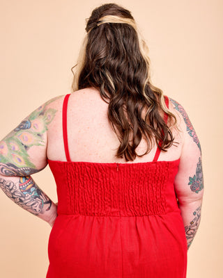 A woman shown from behind wearing a bright red sleeveless smocked dress, with her arms tattooed and her hair styled in loose waves, against a solid beige background. This is the Wayland Dress sewing pattern from Cashmerette.