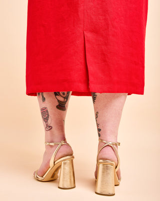A person shown from the back wearing bright red shorts and metallic gold block-heel sandals against a warm beige studio background. This is the Wayland Dress sewing pattern from Cashmerette.