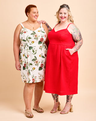 Two women modeling sleeveless midi dresses against a solid beige studio background. The woman on the left wears a white dress with a colorful floral print, and the woman on the right wears a solid bright red dress. This is the Wayland Dress sewing pattern from Cashmerette.