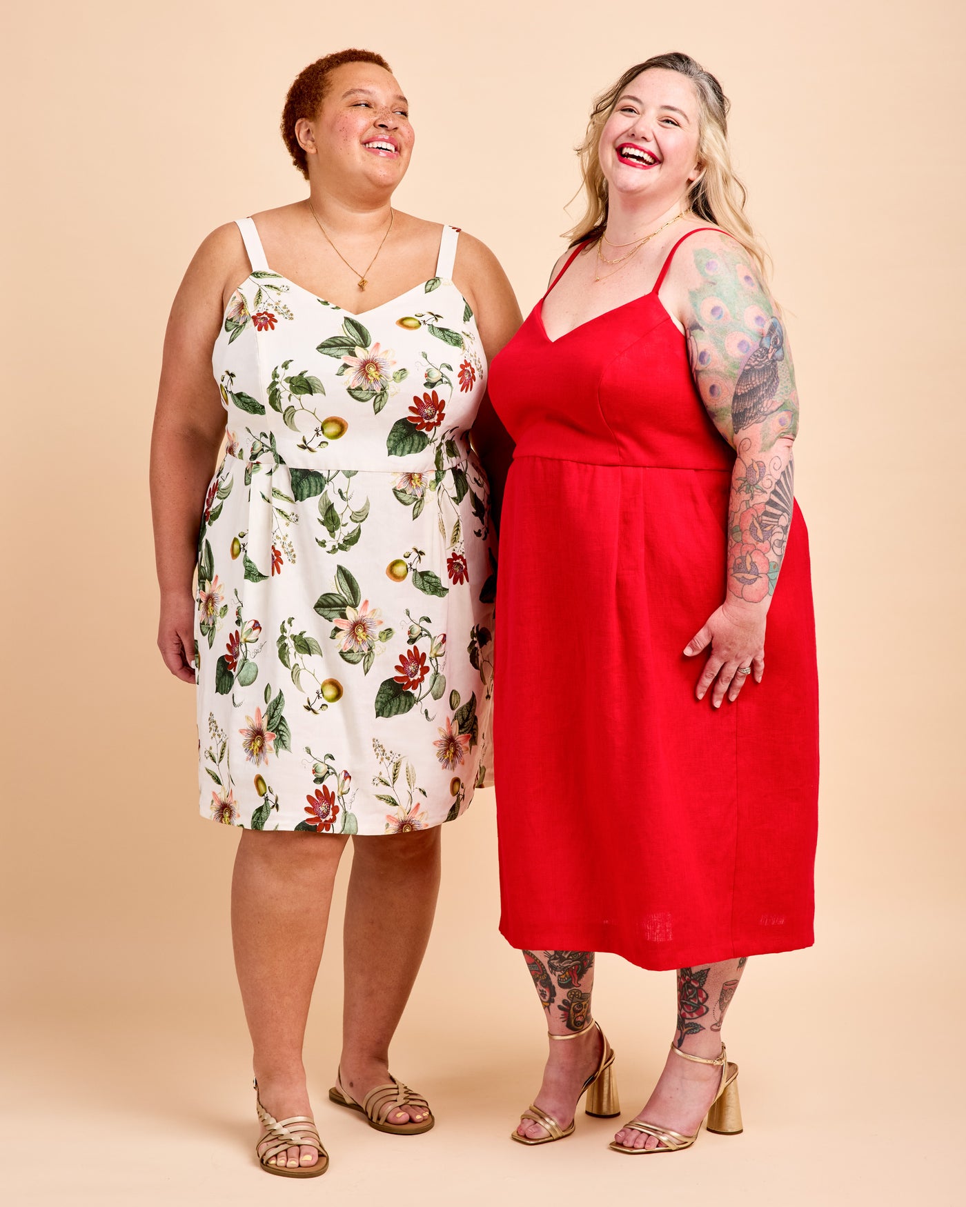 Two women modeling sleeveless midi dresses against a solid beige studio background. The woman on the left wears a white dress with a colorful floral print, and the woman on the right wears a solid bright red dress. This is the Wayland Dress sewing pattern from Cashmerette.