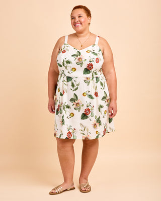 A woman modeling a sleeveless white A-line dress with a green and red floral print, paired with neutral flat sandals, against a solid beige studio background. This is the Wayland Dress sewing pattern from Cashmerette.