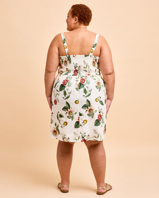 A plus-size model is shown from the back wearing a white sleeveless midi dress with a colorful floral and fruit print, paired with tan flat sandals, against a solid beige background. This is the Wayland Dress sewing pattern from Cashmerette.