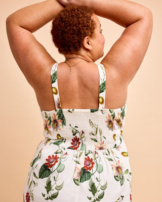 A woman viewed from behind wearing a white sleeveless dress with a colorful floral print and green leafy vines, set against a solid beige background. This is the Wayland Dress sewing pattern from Cashmerette.