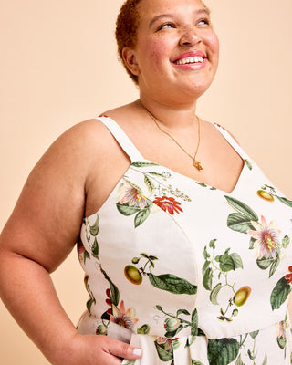 A smiling woman wearing a sleeveless white dress with a colorful botanical print of flowers and green leaves, photographed against a solid beige background. This is the Wayland Dress sewing pattern from Cashmerette.