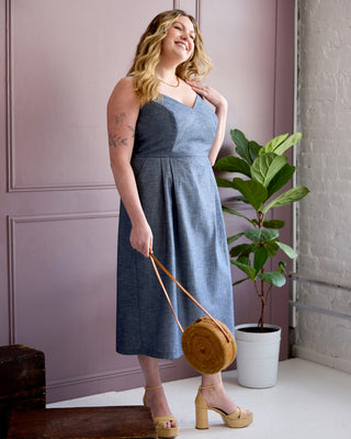 A woman models a sleeveless, V-neck midi dress in a dusty blue denim-like fabric. She is holding a round woven straw handbag with a tan strap and wearing beige platform sandals. This is the Wayland Dress sewing pattern from Cashmerette.