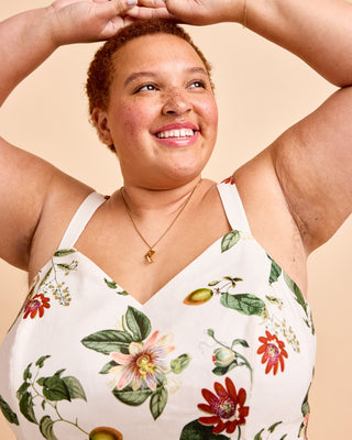 A smiling woman with short auburn hair poses with her arms raised, wearing a white spaghetti-strap top with a colorful botanical print of flowers and leaves, set against a solid beige background. This is the Wayland Dress sewing pattern from Cashmerette.