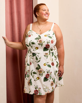 A woman wearing a sleeveless white mini dress with a green and red floral print, standing indoors next to a dusty rose curtain. This is the Wayland Dress sewing pattern from Cashmerette.