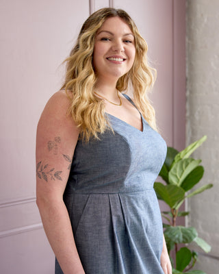 A smiling blonde woman wearing a sleeveless slate blue dress, standing indoors against a pink wall with a green plant in the background. This is the Wayland Dress sewing pattern from Cashmerette.