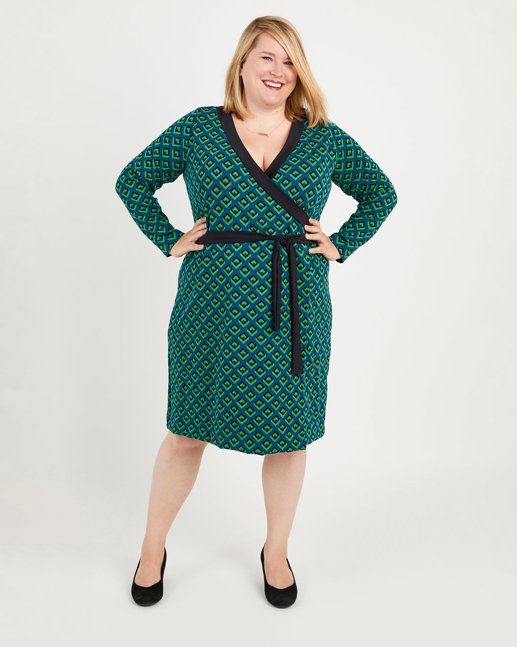 Woman wearing a green patterned dress with a belt on a white background. This dress is the Appleton Wrap Dress sewing pattern from Cashmerette.