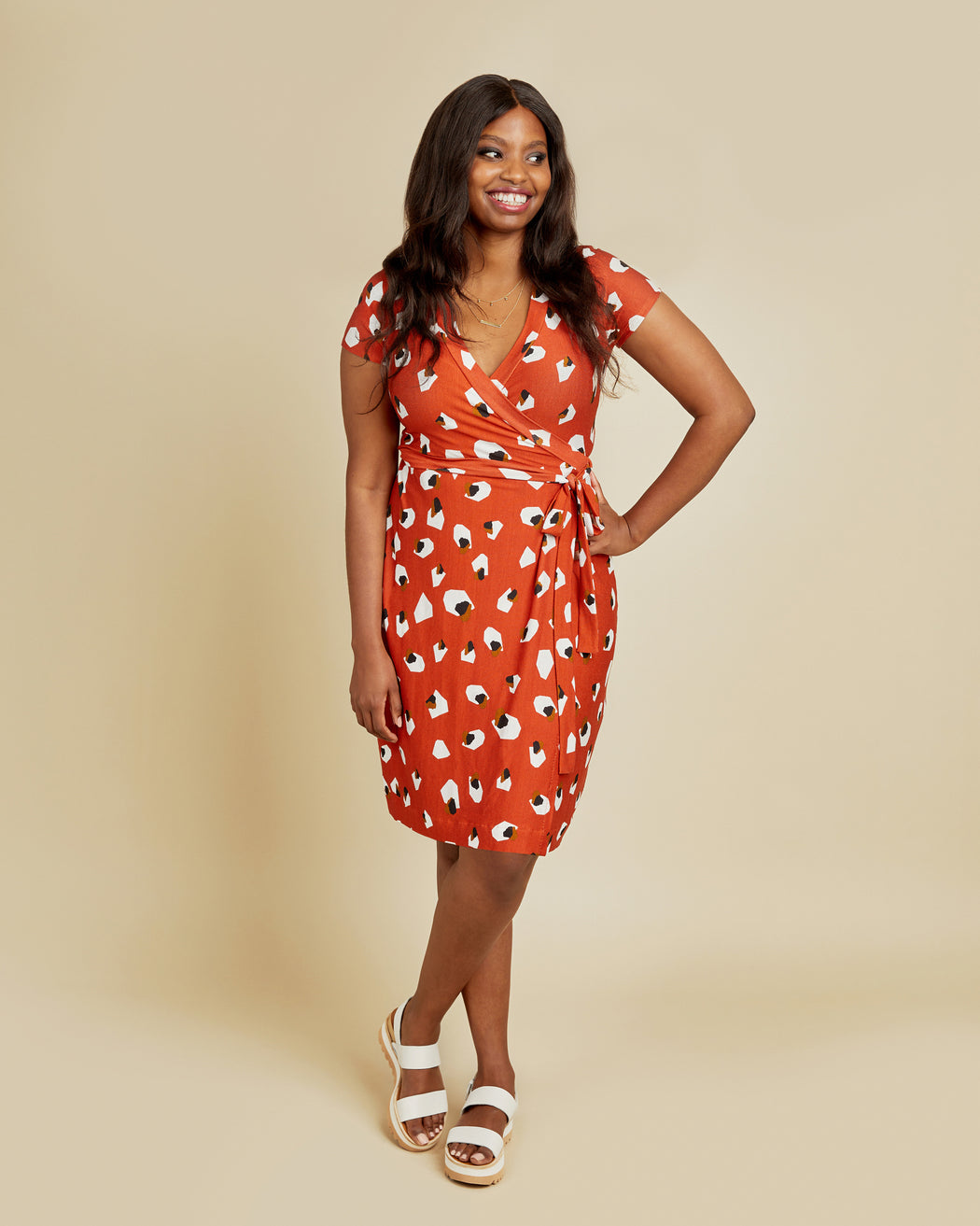 Woman wearing a red dress with white patterns on a beige background. This dress is the Appleton Wrap Dress sewing pattern from Cashmerette.
