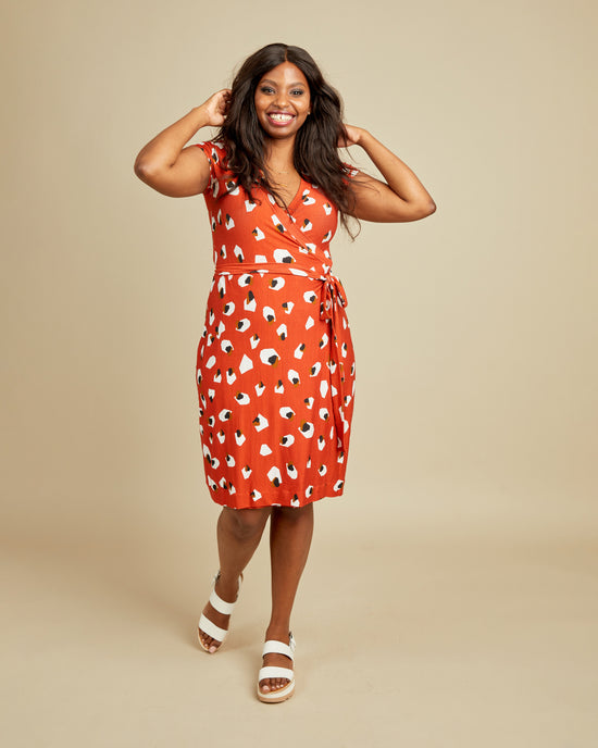 Woman wearing a red dress with a pattern on a beige background. This dress is the Appleton Wrap Dress sewing pattern from Cashmerette.