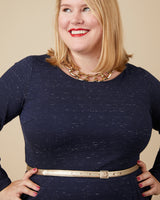 Woman wearing a navy blue dress with a gold belt and necklace against a beige background. This dress is the Rivermont Dress & Top sewing pattern from Cashmerette.