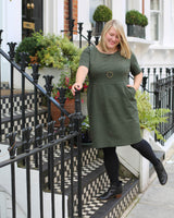 Woman in a green dress standing on a black iron railing with plants and a building in the background. This dress is the Rivermont Dress & Top sewing pattern from Cashmerette.