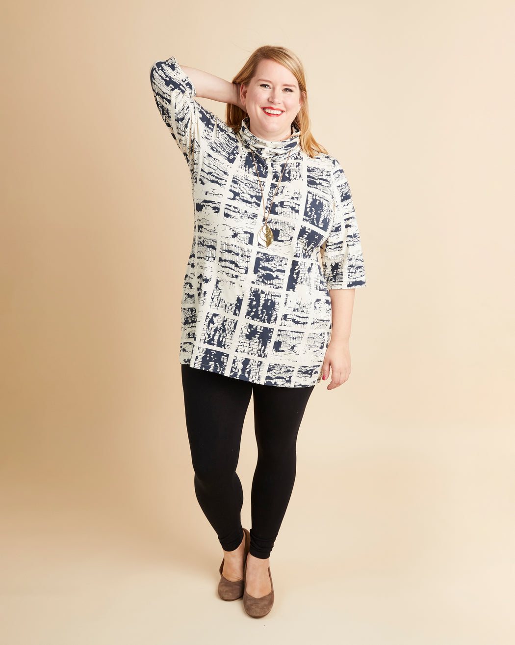 Woman wearing a patterned blouse and black leggings on a beige background. This is the Pembroke Dress & Tunic sewing pattern from Cashmerette.