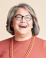 Person with gray hair wearing glasses and a brown shirt with a large orange necklace on a beige background. This is the Grafton Dress, Top & Skirt sewing pattern from Cashmerette.