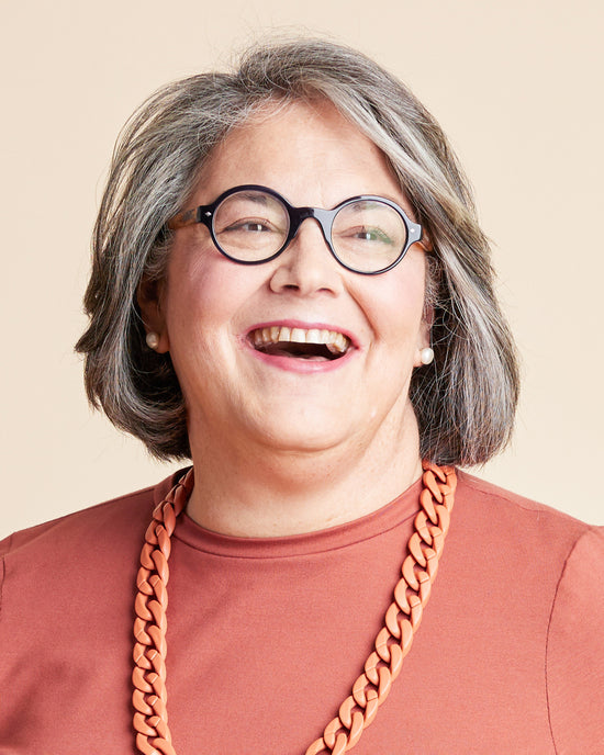 Person with gray hair wearing glasses and a brown shirt with a large orange necklace on a beige background. This is the Grafton Dress, Top & Skirt sewing pattern from Cashmerette.