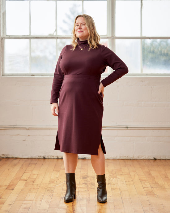 Woman wearing a burgundy dress with a slit standing in a room with large windows.. This is the Grafton Dress, Top & Skirt sewing pattern from Cashmerette.