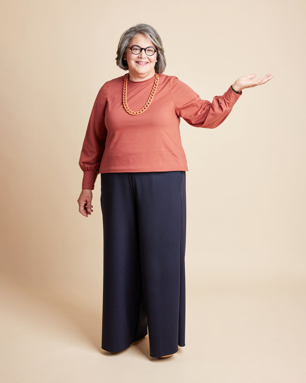 Woman wearing a rust-colored sweater and navy pants on a beige background. This is the Grafton Dress, Top & Skirt sewing pattern from Cashmerette.