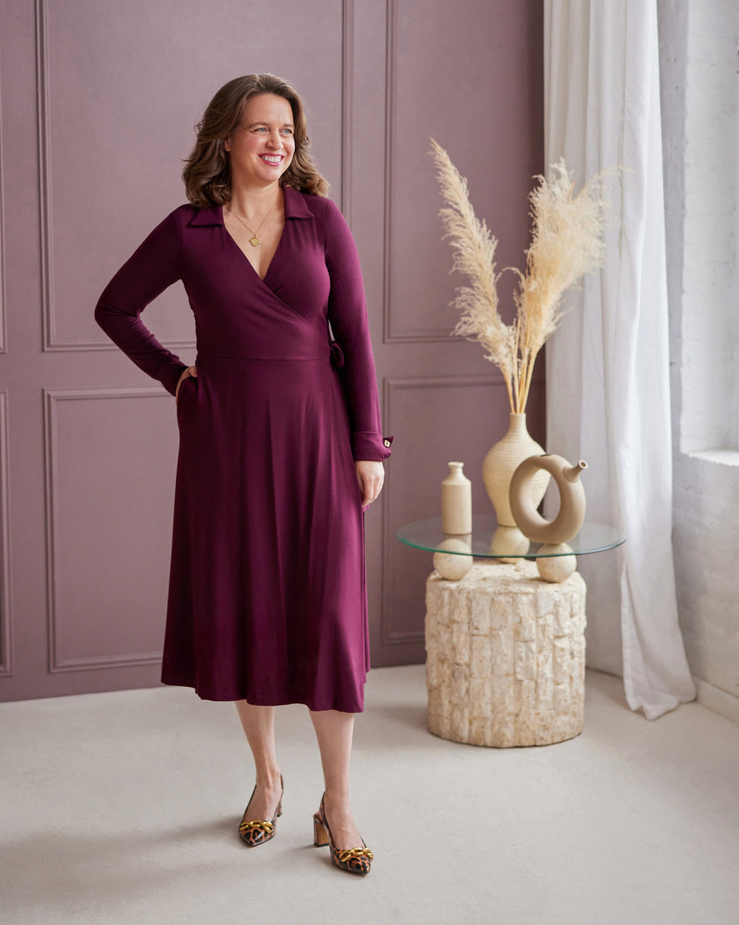 Woman in a purple dress standing in a room with decorative elements. This is the Hazelton Wrap Dress sewing pattern from Cashmerette.