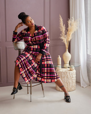 Woman in a plaid dress sitting on a chair in a stylish room with decor elements. This is the Hazelton Wrap Dress sewing pattern from Cashmerette.