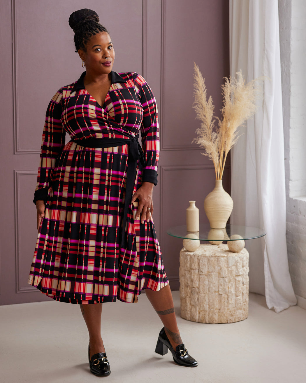 Woman wearing a plaid dress standing in a room with a decorative table and vase. This is the Hazelton Wrap Dress sewing pattern from Cashmerette.