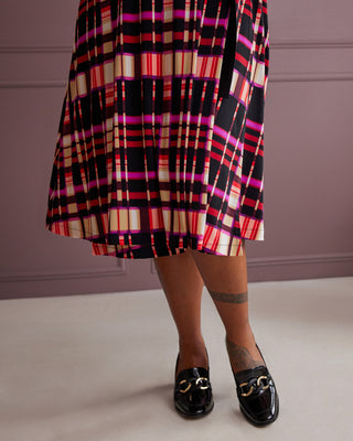 Person wearing a plaid dress with a pattern of red, black, and beige on a plain background. This is the Hazelton Wrap Dress sewing pattern from Cashmerette.