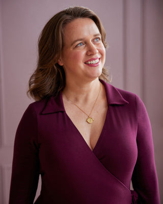 Woman wearing a deep neckline dress with a gold necklace against a pink background. This is the Hazelton Wrap Dress sewing pattern from Cashmerette.