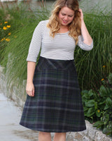 Woman wearing a striped shirt and plaid skirt standing on a sidewalk with plants in the background. This dress is the Washington Dress sewing pattern from Cashmerette.