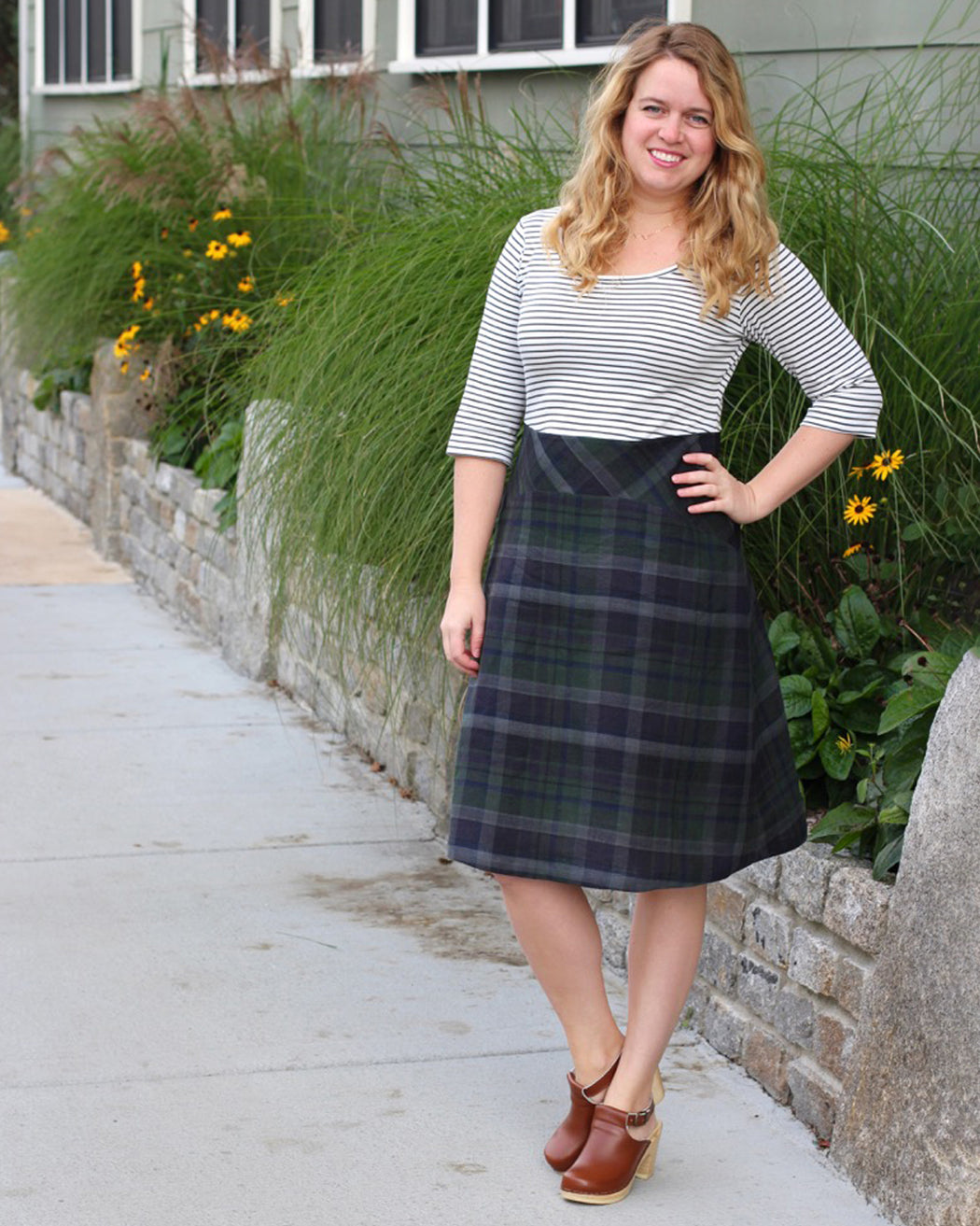 Woman wearing a striped shirt and plaid skirt standing on a sidewalk with plants and flowers in the background. This dress is the Washington Dress sewing pattern from Cashmerette.