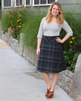 Woman wearing a striped shirt and plaid skirt standing on a sidewalk with plants and flowers in the background. This dress is the Washington Dress sewing pattern from Cashmerette.