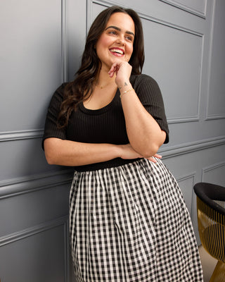 Woman wearing a black top and checkered skirt against a gray paneled wall. This is the Duxbury Dress sewing pattern from Cashmerette.