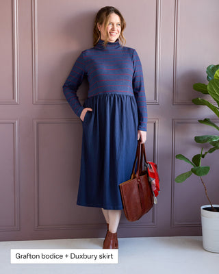 Woman wearing a blue dress with a striped bodice and Duxbury skirt, holding a brown bag against a purple paneled wall. This is the Duxbury Dress sewing pattern from Cashmerette. Over the image is the text "Grafton bodice + Duxbury skirt"