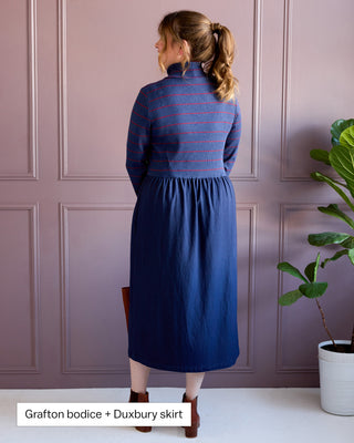 Woman wearing a blue dress with a striped bodice and plain skirt against a pink paneled wall. This is the Duxbury Dress sewing pattern from Cashmerette. Over the image is the text "Grafton bodice + Duxbury skirt"