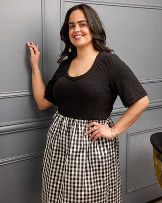 Woman wearing a black top and checkered pants standing against a gray wall. This is the Duxbury Dress sewing pattern from Cashmerette.