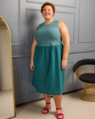 Woman wearing a teal dress standing in a room with gray walls and a chair. This is the Duxbury Dress sewing pattern from Cashmerette.