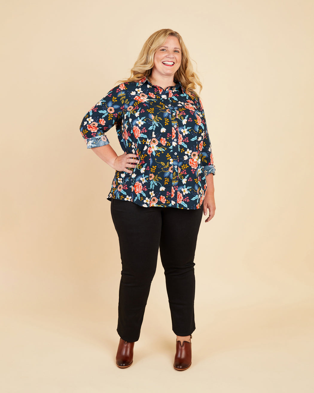 Woman wearing a floral blouse and black pants on a beige background. This button-up shirt is the Harrison Shirt sewing pattern from Cashmerette.