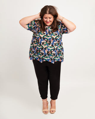 Woman wearing a floral blouse and black pants on a plain background. This top is the Montrose Top sewing pattern from Cashmerette.