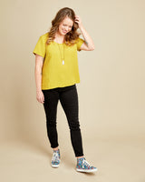 Woman wearing a yellow top and black pants with colorful shoes on a beige background. This top is the Montrose Top sewing pattern from Cashmerette.