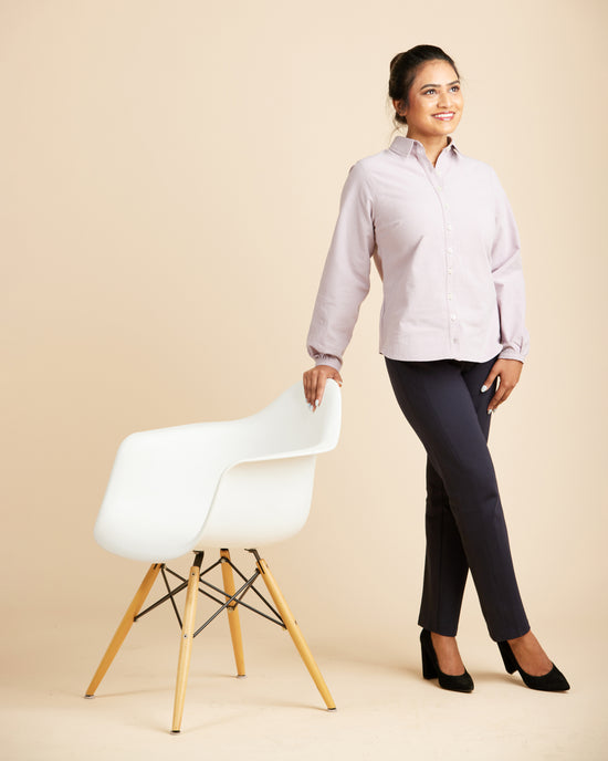 Woman in a light purple shirt and dark pants standing next to a white chair on a beige background. This top is the Vernon Shirt sewing pattern from Cashmerette.