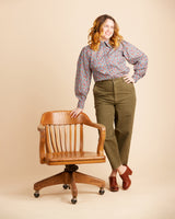 Woman in a patterned blouse and green pants standing next to a wooden chair on a beige background. This top is the Vernon Shirt sewing pattern from Cashmerette.