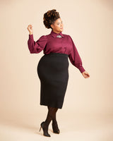Woman wearing a burgundy blouse and black skirt on a beige background. This top is the Vernon Shirt sewing pattern from Cashmerette.