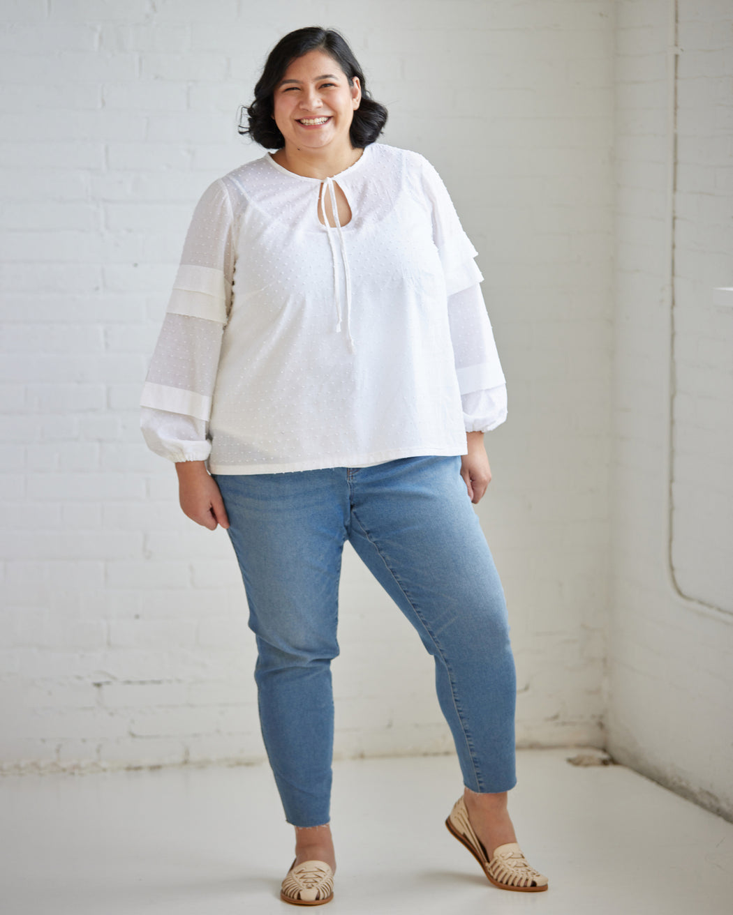 Woman wearing a white blouse and blue jeans standing against a white brick wall. This top is the Alton Blouse sewing pattern from Cashmerette.