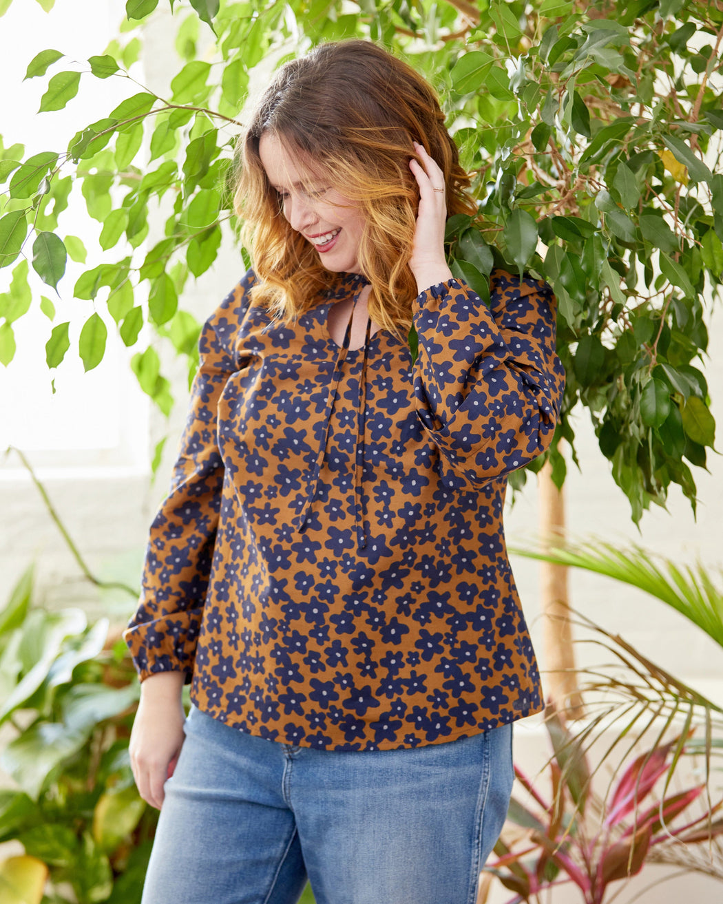 Woman wearing a floral blouse and jeans standing among plants. This top is the Alton Blouse sewing pattern from Cashmerette.