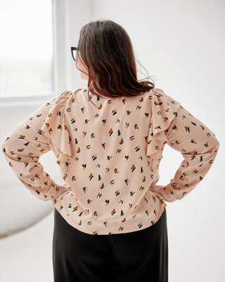 Woman wearing a beige blouse with a subtle pattern, standing against a white wall. This top is the Selwyn Top sewing pattern from Cashmerette.