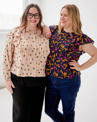 Two women standing together, one wearing a beige blouse with small patterns and black pants, the other in a colorful patterned top and blue jeans. This top is the Selwyn Top sewing pattern from Cashmerette.