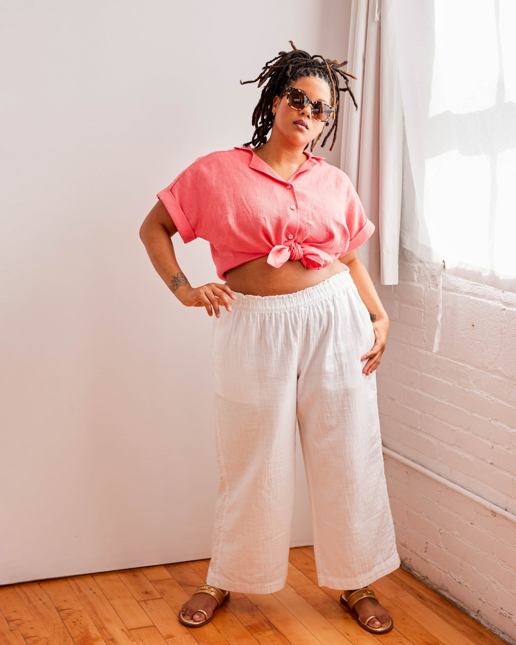 Person wearing a pink top and white pants standing in a room with a window. Woman wearing a plaid shirt and denim shorts against a neutral background. This top is the Haskell Shirt sewing pattern from Cashmerette.