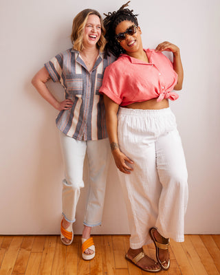 Two women standing together against a plain wall on a wooden floor. Woman wearing a plaid shirt and denim shorts against a neutral background. This top is the Haskell Shirt sewing pattern from Cashmerette.