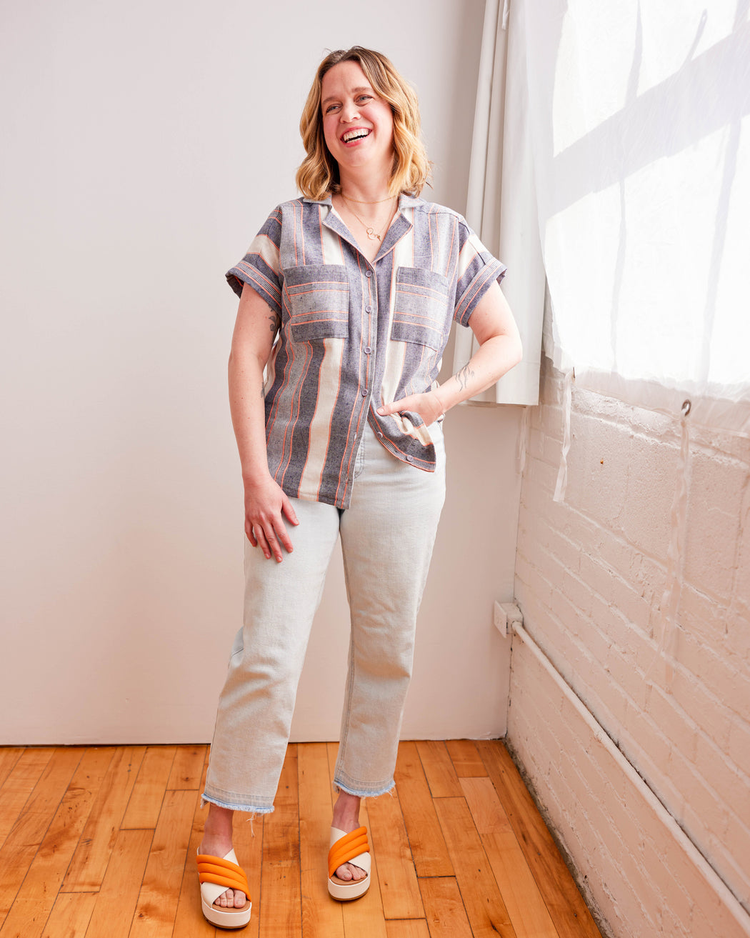 Woman standing in a room with wooden floor and white walls, wearing a plaid shirt and white pants. Woman wearing a plaid shirt and denim shorts against a neutral background. This top is the Haskell Shirt sewing pattern from Cashmerette.